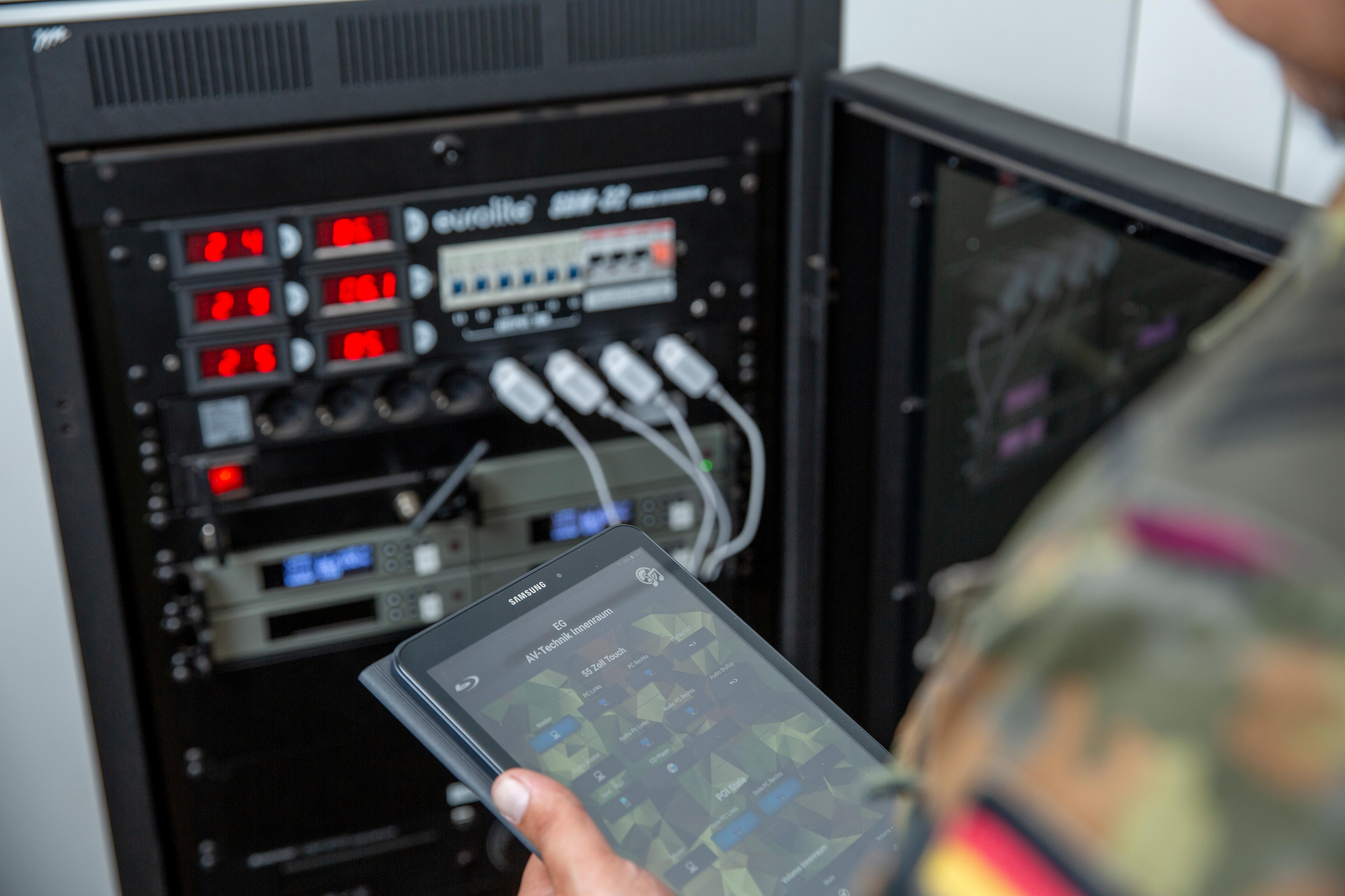 Eine große Technikanlage mit verschiedenen Anzeigen und Kabeln. Davor steht ein Soldat mit einem Tablet.