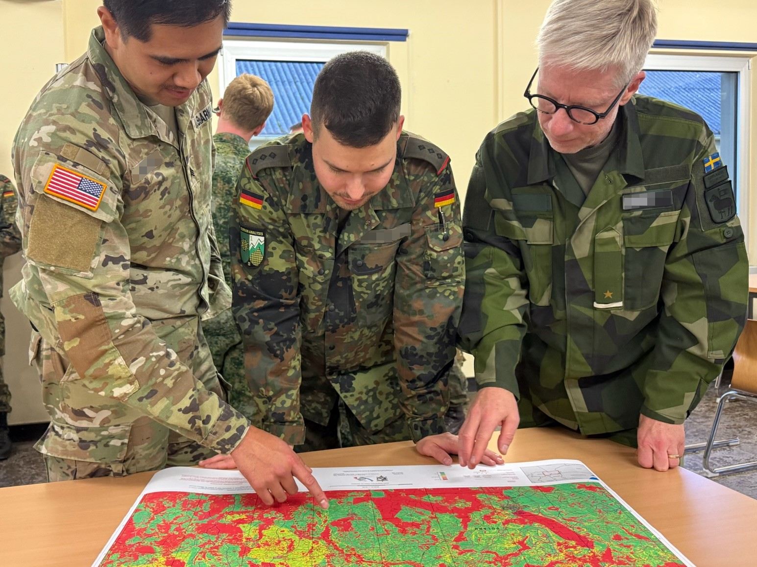 Drei Soldaten aus unterschiedlichen Ländern schauen gemeinsam auf eine Karte mit Geländedaten auf einem Tisch.