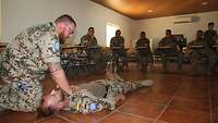 Soldaten sitzen im Unterrichtsraum. Der Ausbilder kniet neben dem Übungsverletzten und zeigt die Erste-Hilfe-Maßnahmen