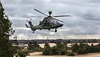 Kampfhubschrauber Tiger fliegt bei bewölktem Himmel über sandiger Landschaft mit grünen Sträuchern.