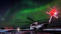 Ein Hubschrauber CH-53 steht auf einem leicht beschneiten Flugfeld. Am Himmel leuchtet grünes Polarlicht.