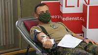 Ein Soldat mit Mund-Nasenschutz liegt auf einer Liege. In seinem gestreckten Arm steckt eine Nadel, daran hängt ein Schlauch.