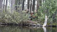 Zwei Soldaten knien nahe einem Fluss. Einer von beiden sieht durch das Visier der Waffe