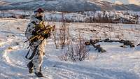 Ein Soldat in Schneetarnuniform geht in verschneiter Landschaft mit einem Gewehr in der Hand einen Landweg entlang.