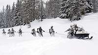 Ein Ski-Doo zieht eine Gruppe Soldaten auf Skiern an einer Leine durch den Schnee hinter sich her.