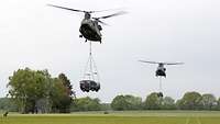 Zwei Hubschrauber transportieren zwei Einsatzfahrzeuge Spezialisierte Kräfte Mungo an einer Halterung hängend über eine Wiese.