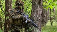 Ein getarnter Soldat kniet in einem Wald an einem Baum und hält das Gewehr G36