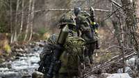 An einem steinigen Bach laufen zwei Bundeswehrsoldaten mit einer Panzerfaust auf dem Rücken in ihre Stellungen.