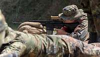 Ein Soldat zielt mit einem auf Sandsäcken aufgelegten Gewehr auf das vor ihm liegende Gelände.