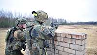 Ein Soldat steht hinter einem Mauerrest und zielt auf das Ziel. Ein weiterer Soldat beaufsichtigt den Schützen.