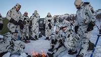 Soldaten in Schneeflecktarn stehen und sitzen um eine Feuerstelle im Schnee.