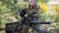 Ein Soldat sitzt im Gras hinter einer Granatmaschinenwaffe und öffnet die Verschlussklappe der Waffe