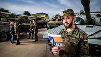 Ein Soldat steht mit einem Flyer in der Hand auf einem Gelände mit mehreren Bundeswehrfahrzeugen und schaut sich um.