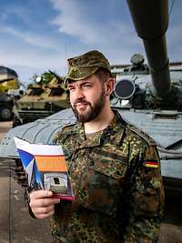 Ein Soldat steht mit einem Flyer in der Hand auf einem Gelände mit mehreren Bundeswehrfahrzeugen und schaut sich um.