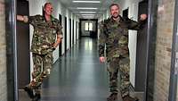 Zwei Soldaten in zwei verschiedenen Flecktarn-Uniformen stehen in einem langen Gang an Türen, die sich gegenüberliegen.