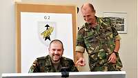 Ein Soldat sitzt vor einer großen Tafel, ein anderer steht daneben und zeigt auf die Tafel, beide lächeln.
