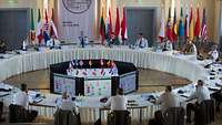 Zahlreiche Generale sitzen an einem runden, weiß eingedeckten Tisch, hinter ihnen die Flaggen der Teilnehmerländer.