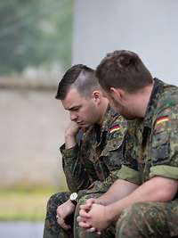Zwei Soldaten sitzten leicht gekrümmt vor einem Gebäude auf einer Holzbank und unterhalten sich