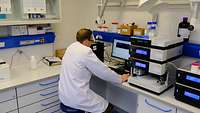 Ein Mann sitzt in einem Labor vor einem Bildschirm, neben ihm stehen Diagnosegeräte