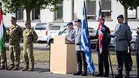 Mehrere Soldaten stehen neben einem Pult, zwei halten Flaggen in den Händen. Ein Soldat steht an einem Pult und hält eine Rede.