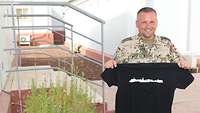 Neben einem Pflanzenkübel steht ein Soldat und hält ein T Shirt mit der Silhouette von Kassel in den Händen