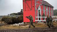 Fünf bewaffnete Soldaten laufen über ein Gefechtsfeld, im Hintergrund eine rote Gebäudeattrappe.
