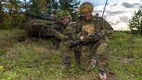 Zwei getarnte und bewaffnete Soldaten hocken an einem Waldrand, hinter ihnen steht ein Panzer.