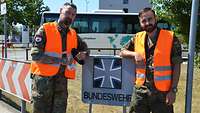 Zwei Soldaten vor einem kleinen Bundeswehr-Schild, im Hintergrund ein Reisebus. 