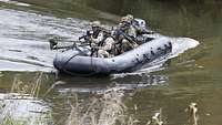 Vier Soldaten sitzen bewaffnet in einem Schlauchboot und fahren über einen Fluss Richtung Ufer.