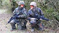 Zwei kniende und bewaffnete Soldaten sprechen an einem Waldrand über die aktuelle Gefechtslage.