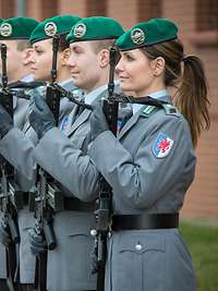 Bewaffnete Soldatinnen und Soldaten im Dienstanzug mit Waffen sind in einer Reihe angetreten.