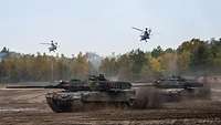 Panzer fahren über ein Feld, darüber kreisen zwei Hubschrauber