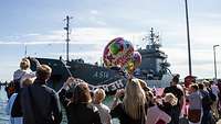 Ein Schiff läuft in einen Hafen ein. Dort stehen wartende Menschen.