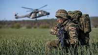 Ein bewaffneter Soldat kniet in einem Getreidefeld. Er sichert die Landung eines Hubschraubers.