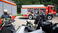 Zwei Feuerwehrmänner stehen an einem Unfall-Fahrzeug, im Hintergrund zwei Bundeswehr-Feuerwehrfahrzeuge.