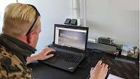 Ein Soldat sitzt vor einem Laptop, auf dessen Bildschirm das Kopfteil einer Gesichtsschutzmaske abgebildet ist