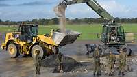 Zwei Bagger und mehrere Soldaten mit Schaufeln reparieren einen Krater auf dem Flugplatz.