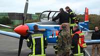 Soldaten und Feuerwehrleute stehen an dem blau-rot-weißen Propeller-Flugzeug Pilatus PC-9, dessen Cockpit aufgeklappt ist.