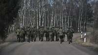 Eine Gruppe von Rekrutinnen und Rekruten marschiert mit Rucksack und im Laufschritt auf einer Straße in Richtung Wald.