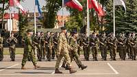 Drei Offiziere marschieren im Gleichschritt vor einer Front bewaffneter Soldaten. Dahinter sind Flaggen vieler Nationen zu sehen