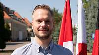 Veit Rauen im Portrait vor einer polnischen, dänischen und deutschen Flagge. Im Hintergrund ein Kasernengebäude .