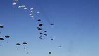 Dutzende Soldaten schweben unter blauem Himmel mit ihren geöffneten Fallschirmen zu Boden.