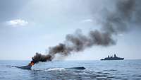 Auf einer Wasseroberfläche brennt ein fast versunkenes Boot, im Hintergrund ein Kriegsschiff.