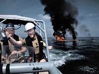 Auf See brennen Reste eines Bootes, im Vordergrund deutsche Marinesoldaten in beigefarbener Arbeitsuniform.
