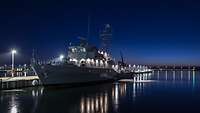 Nachts, ein kleines graues Schiff liegt im Hafen an einer Pier, im glatten Wasser spiegeln sich die Lichter des Schiffs.