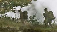 Drei Soldaten laufen mit ihrem Marschgepäck an Rande eines Gefechtsfeldes. Rauch steigt auf.