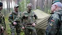 Ein General und drei Soldaten stehen im Wald vor Zelten und unterhalten sich.