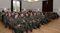 Mehrere Soldaten und Zivilisten sitzen in einem großen Saal zusammen.