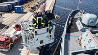 Zwei Feuerwehrmänner stehen im Rettungskorb eines Feuerwehrfahrzeuges, unter ihnen ein Schiff der deutschen Marine.