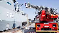 Ein Hubrettungsfahrzeug der Marine mit ausgefahrener Leiter steht neben einem Schiff.